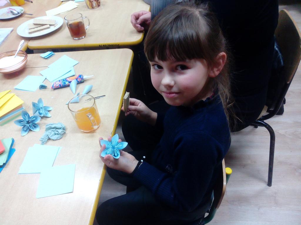 Dziewczynka siedzi przy stole, w lewej ręce trzyma wykonany z papieru niebieski kwiat